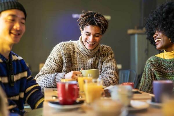 Three students wearing cozy sweaters share a laugh and refreshments around a table. The focus is on a smiling student with textured hair, holding a mug. Various mugs and glasses of orange juice are on the table.