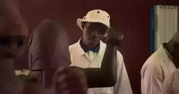 A man in a white lab coat and bucket hat examines the arm of a bronze, Benin sculpture. Other individuals in lab coats are visible in the background. Another sculpture is slightly blurred in the foreground.