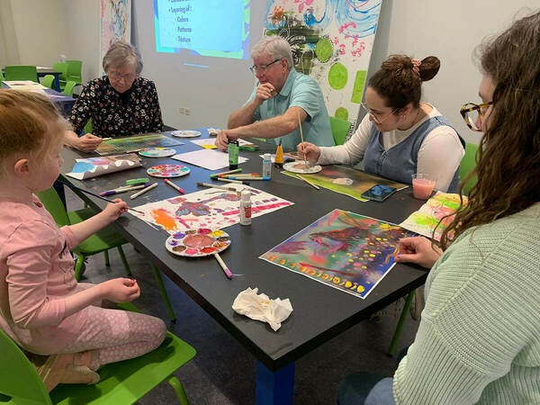 A group of people, including a young child and older adults, sit around a table painting.  They use brushes and palettes with various colors.  A projected presentation on layering with color, pattern, and texture is visible in the background.