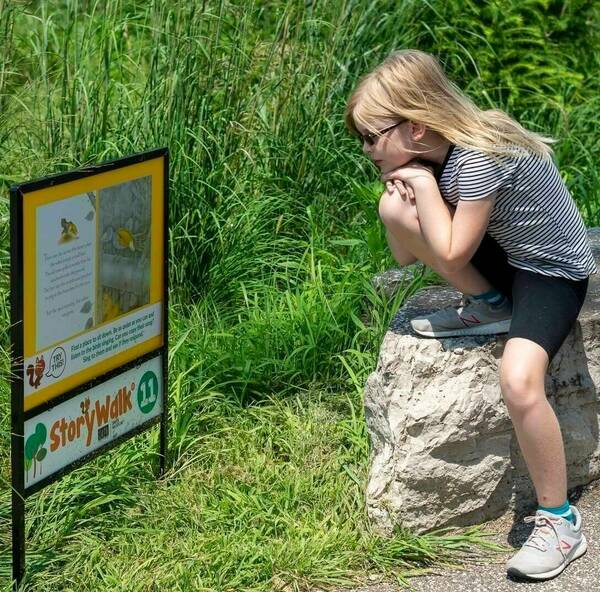 A blonde girl wearing a striped shirt and sunglasses sits on a rock, looking thoughtfully towards a StoryWalk® display surrounded by tall grass.