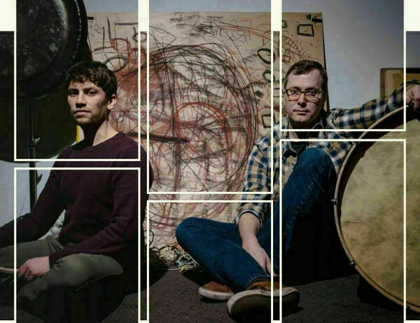Two men sit on a dark floor near a large artwork of red and black scribbles and a large circular drum.  One man wears a dark purple sweater and olive green pants; the other, glasses, a plaid shirt, and jeans.