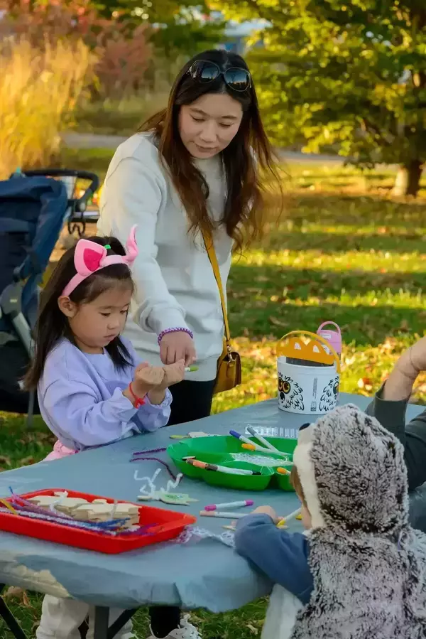 A woman helps a young girl wearing pink animal ears with a craft project at an outdoor table.  Another child in a fuzzy animal costume works on a craft at the other side of the table.  Craft supplies and an owl-decorated bucket are visible.