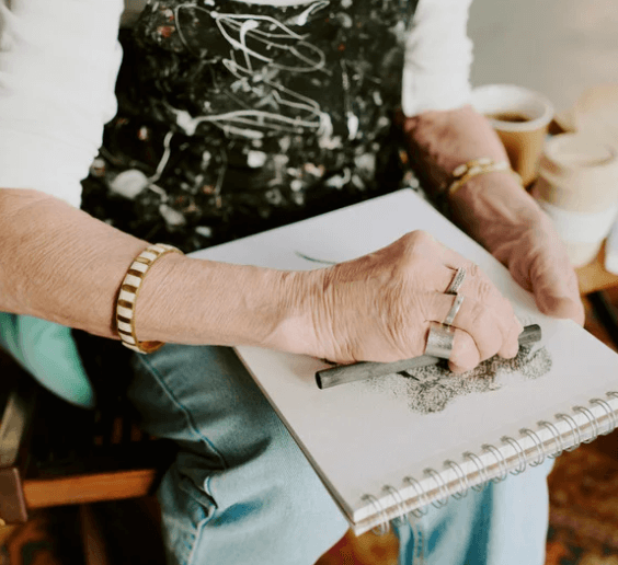 An artist wearing a paint-splattered apron uses a charcoal stick to create a drawing on a spiral-bound sketchbook.