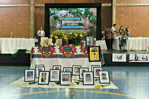 A colorful ofrenda, or altar, honoring victims, with framed photos, flowers, and candles. A large screen displays a banner for the 55th anniversary of the massacre of El Mozote in El Salvador.  A woman stands at a podium and a small group gathers at the side of the stage.