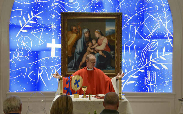 A priest in red vestments leads a small congregation in prayer. He stands at an altar beneath a framed painting of the Holy Family and before a vibrant blue stained-glass window featuring white symbolic imagery.
