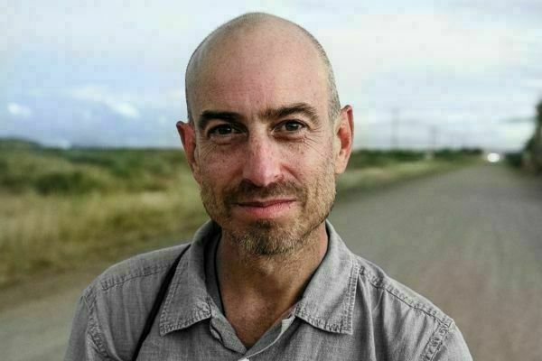 Bald man with a short beard wearing a grey shirt, looking at the camera with a slight smile. Behind him is a blurry dirt road stretching into the distance and a grassy landscape under an overcast sky.