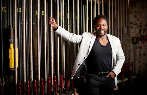 A smiling Black man in a gray blazer and black shirt leans against vertical ropes of a theater fly system, his left hand in his pocket.