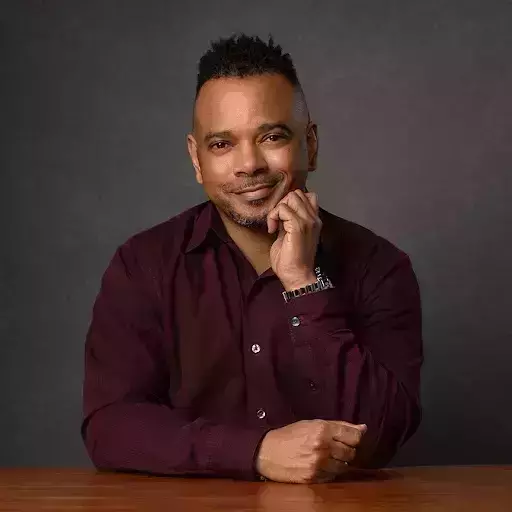 A dark-skinned man with textured hair and a maroon shirt smiles, resting his left hand on his chin. He is seated at a wooden table against a dark gray background.