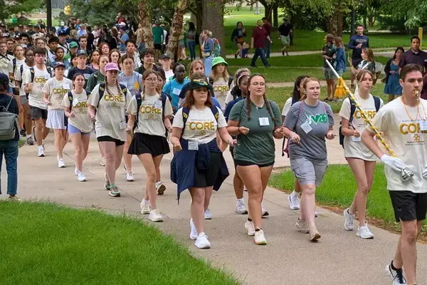 Members of the Notre Dame marching band parade down a campus sidewalk in neat rows, led by 2025 drum major Sean Guibord. Groups of onlookers watch and take photos.