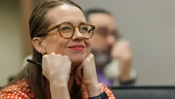 Smiling woman with brown hair, tortoiseshell glasses, and a red polka dot shirt, chin resting on clasped hands, holding a pen.