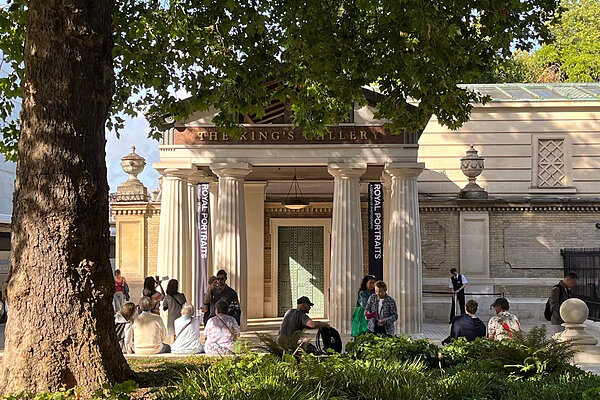 The King's Gallery building with white columns and "ROYAL PORTRAITS" banners. Visitors sit under a large tree.